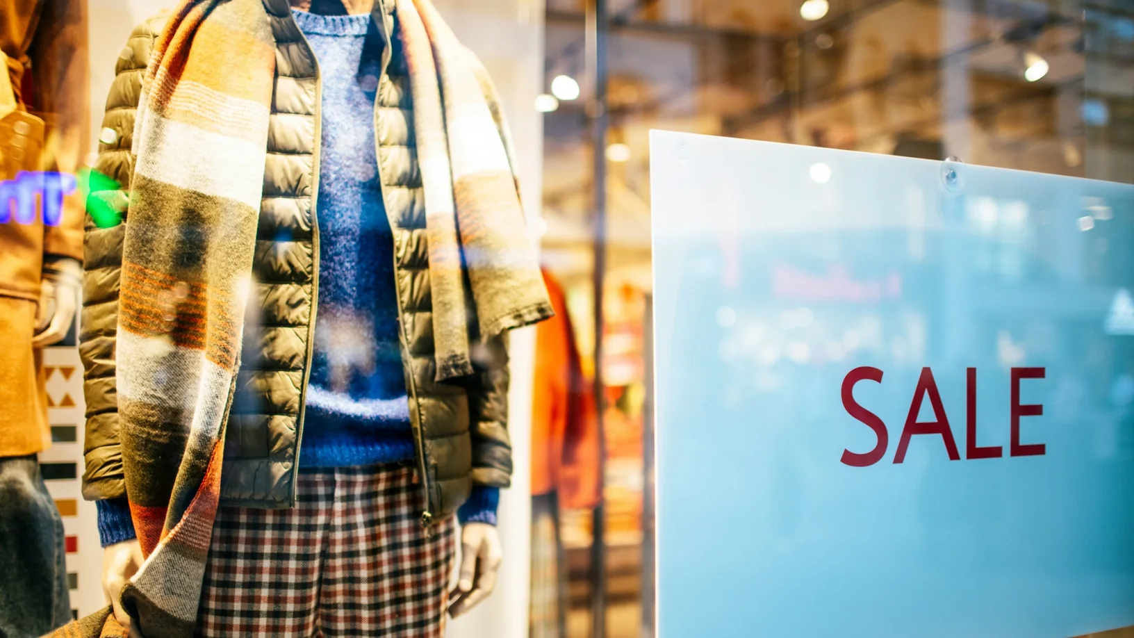 Maniquí con plumífero, bufanda de rayas y pantalón de cuadros en un escaparate, junto a un cartel de rebajas.