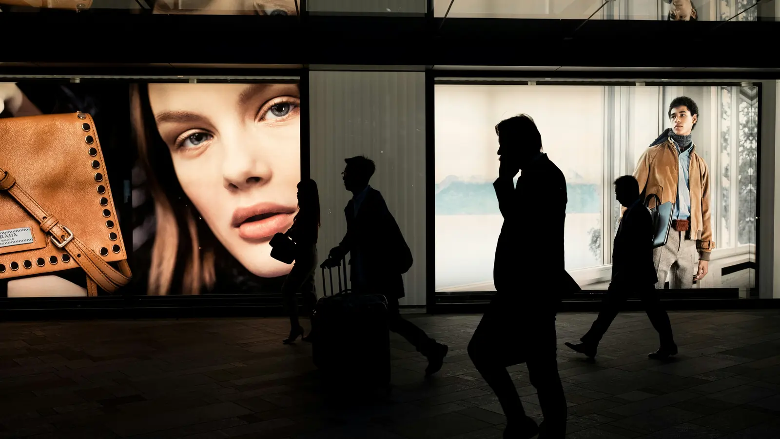 Siluetas de tres personas caminando por un corredor comercial frente a grandes paneles publicitarios retroiluminados.